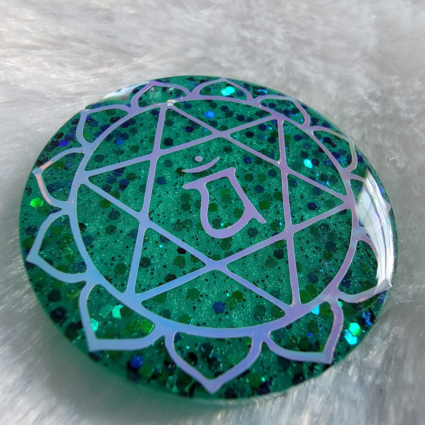 Round epoxy resin piece made with sparkling green glitter and pigment, has heart chakra symbol made with opal white vinyl.