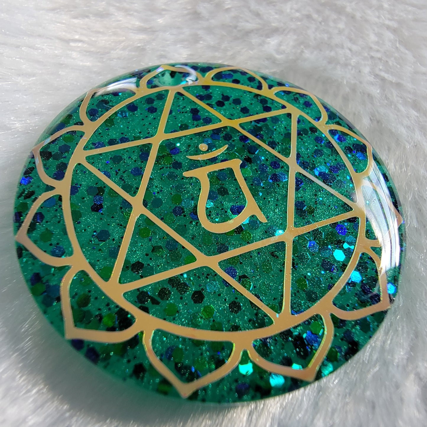 Round epoxy resin piece made with sparkling green glitter and pigment, has heart chakra symbol made with golden vinyl.