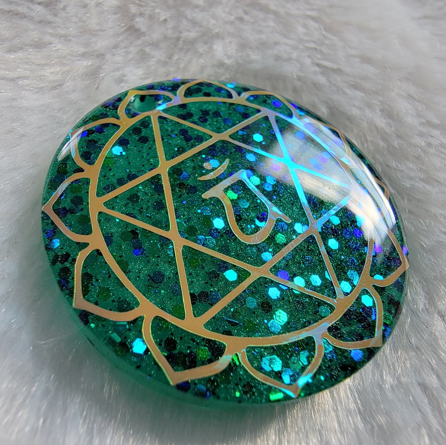 Round epoxy resin piece made with sparkling green glitter and pigment, has heart chakra symbol made with golden vinyl.