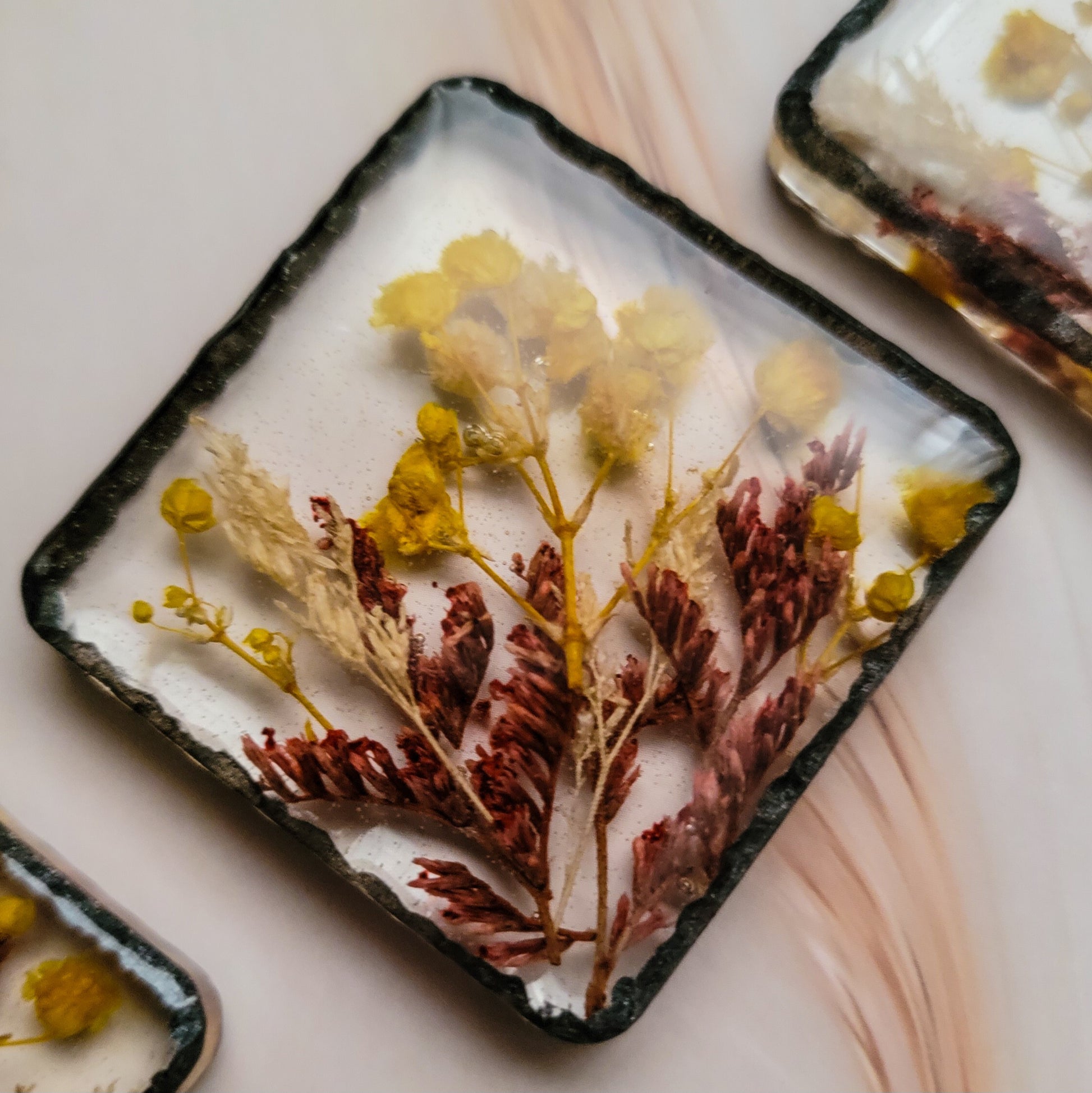 Square epoxy resin piece with yellow, cream and red flowers cast in it. Edges have a faux hammered metal effect with dark metallic black pigment.