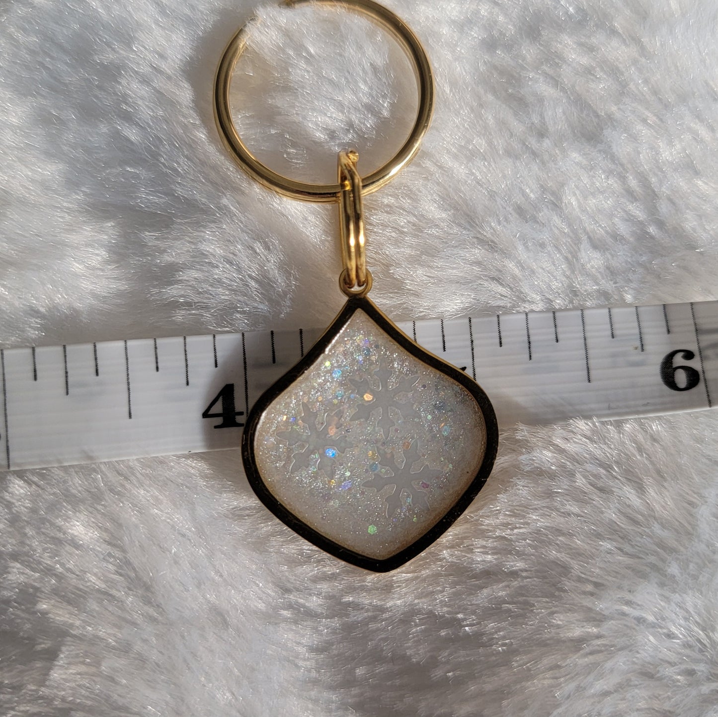 Small metal charm with white glittery snowflake epoxy resin filler, shown with tape measure for size reference. 