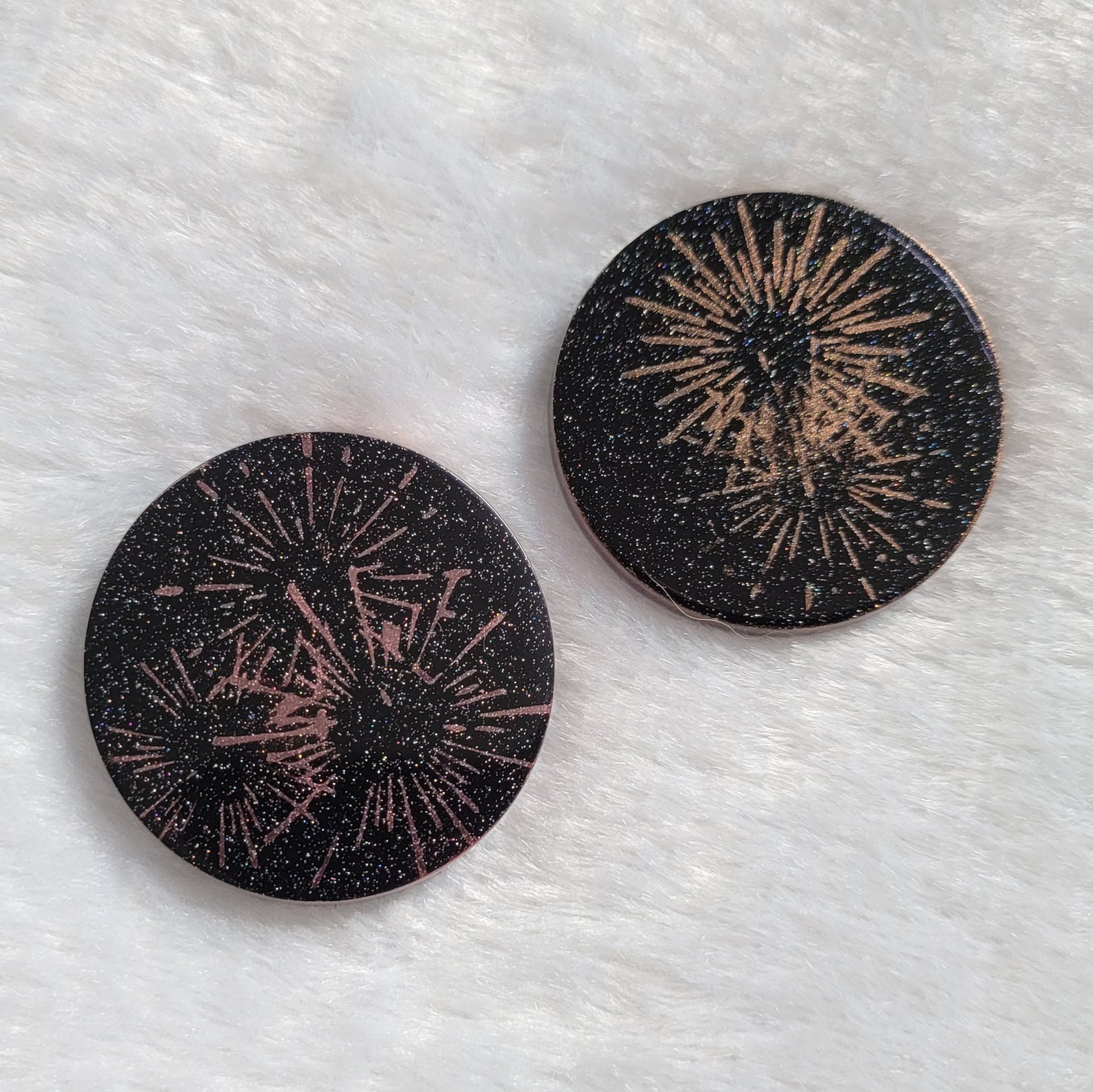 Two round epoxy resin pieces, translucent black with holographic sparkle and metallic fireworks design.