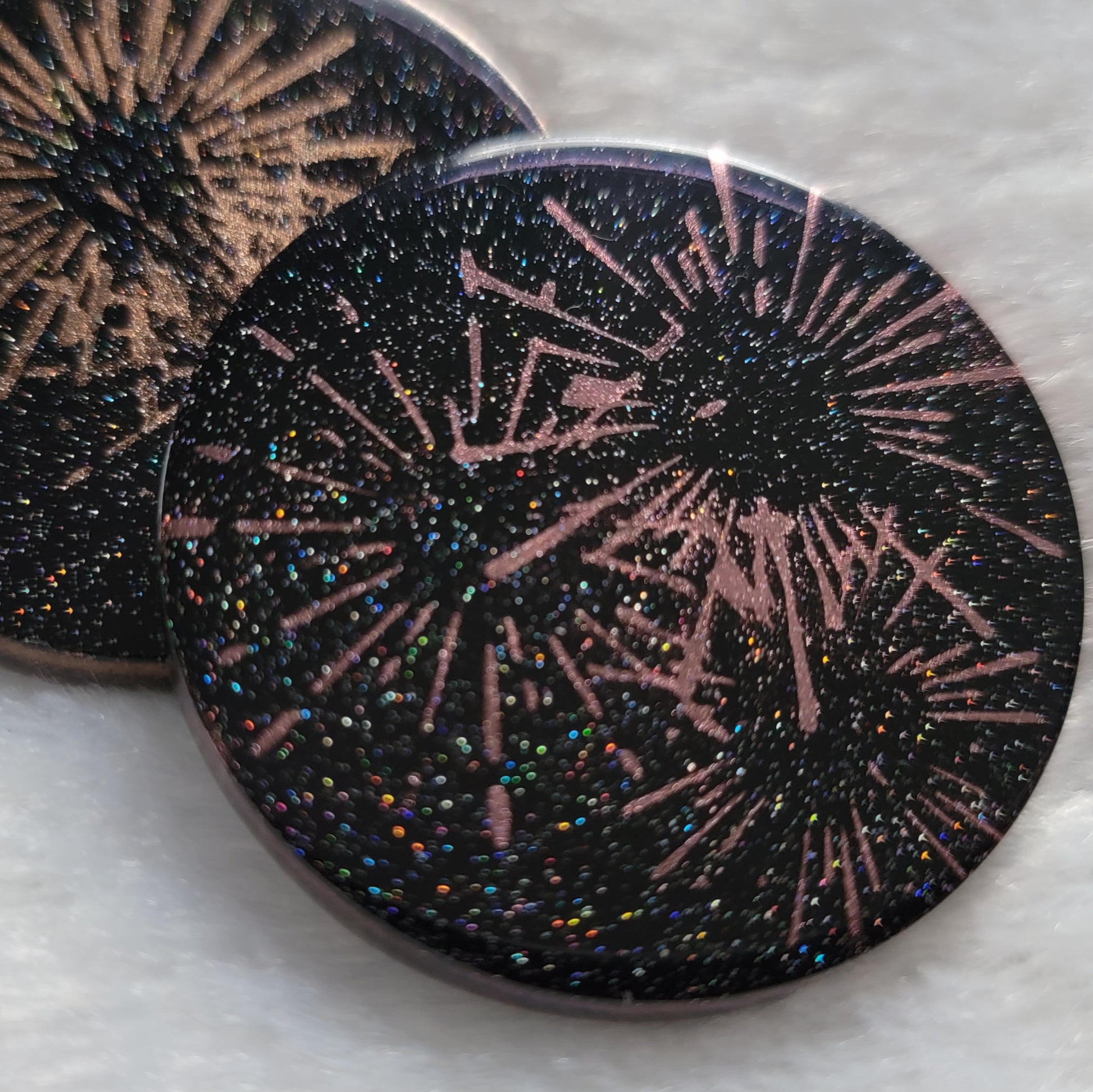 Round epoxy resin piece, translucent black with holographic sparkle and metallic fireworks design.