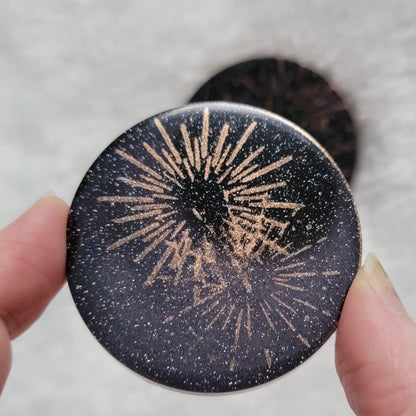 Round epoxy resin piece, translucent black with holographic sparkle and metallic fireworks design. Held up to show translucent effect.