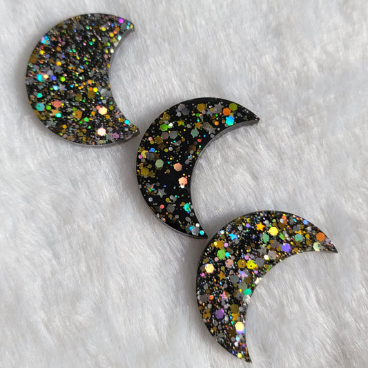 Three crescent moon shaped epoxy resin pieces, black with silver and gold holographic glitter.