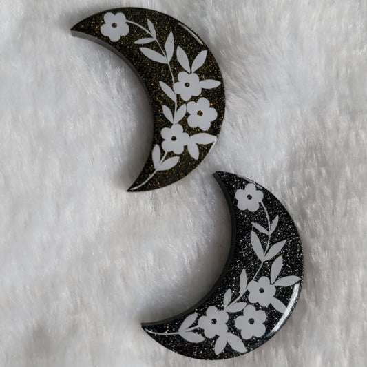 Two crescent moon shaped epoxy resin pieces, black with silver and gold holographic sparkle and floral design in white vinyl.