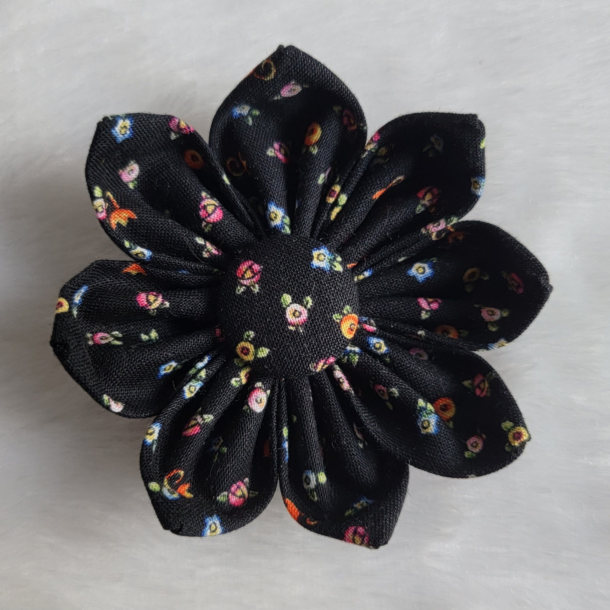 Fabric flower with eight petals and fabric covered button center, print is black with small brightly colored blossoms.