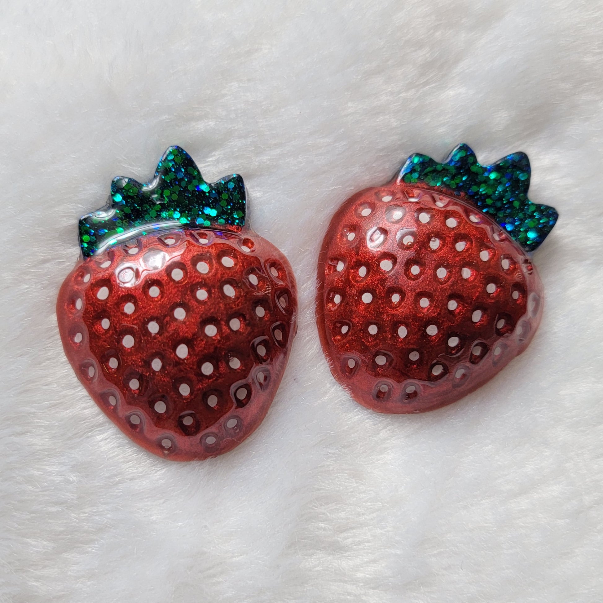 Two strawberry shaped epoxy resin pieces, bright metallic red pigment with hand painted white seeds and leaves in green glitter.