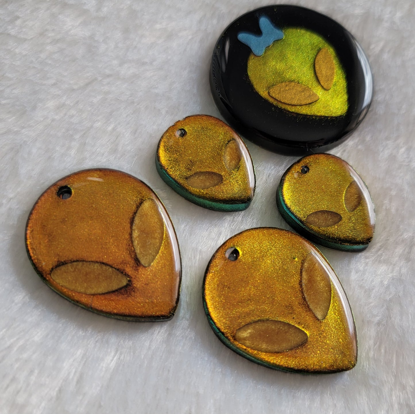 Alien head shaped epoxy resin pieces in iridescent gold, and round black epoxy resin piece with iridescent gold alien head with blue bow.