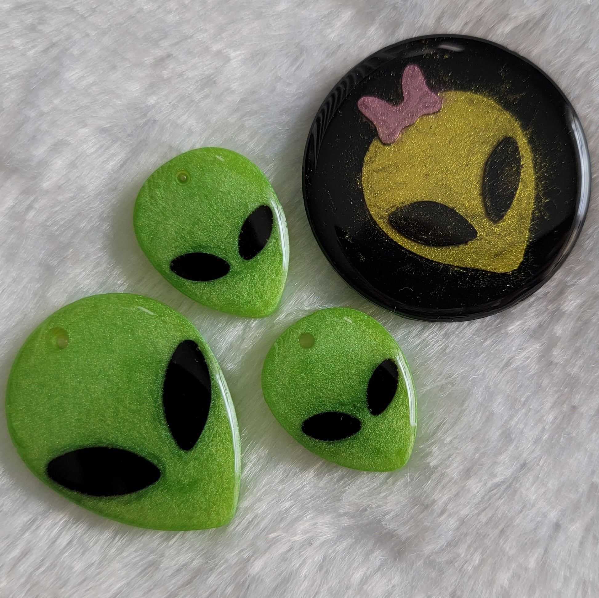 Green alien head shaped epoxy resin piece, and round black epoxy resin piece with green alien head with pink bow.