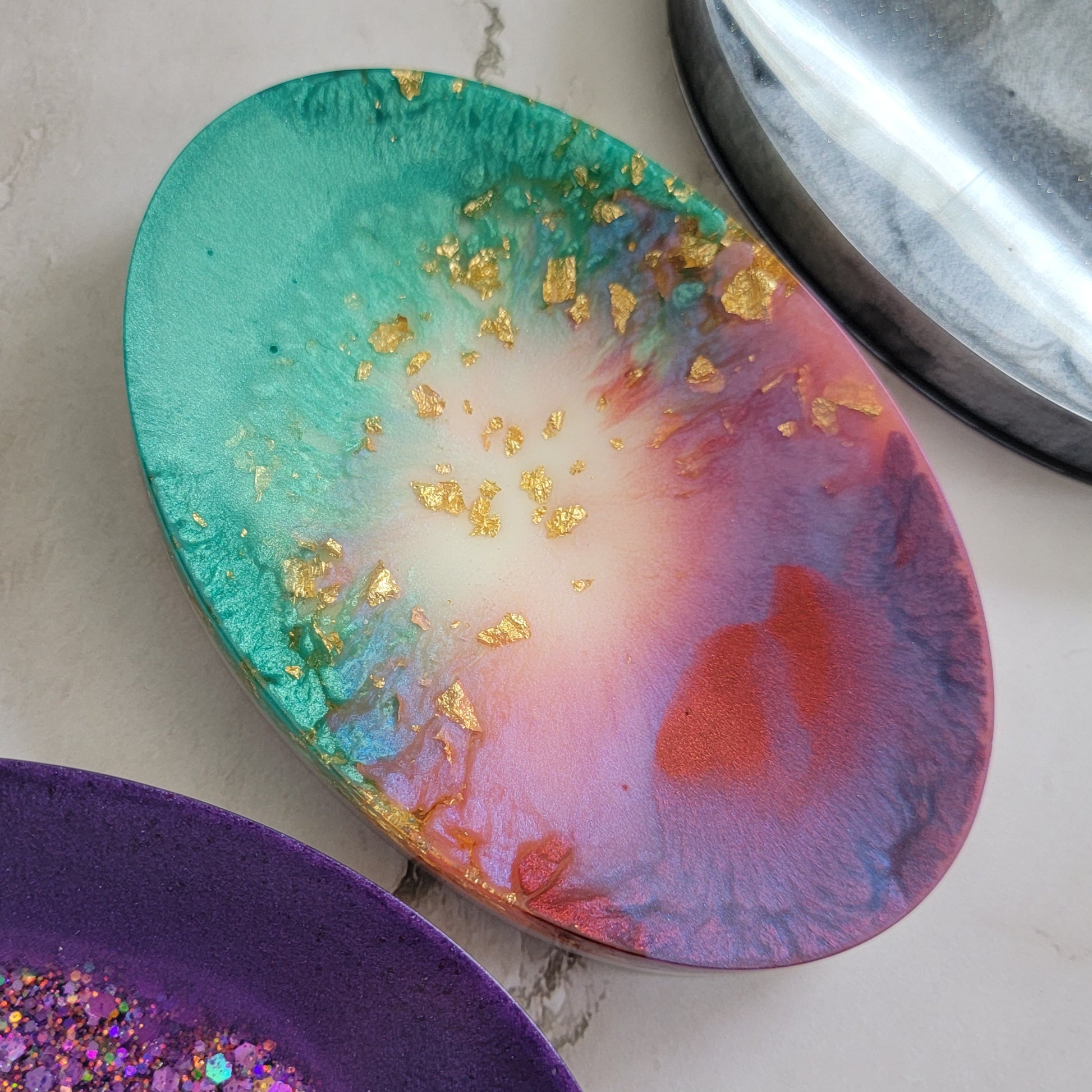 Round epoxy resin soap dish with gold foil flake and swirls of colors including aqua, pink, red and white.