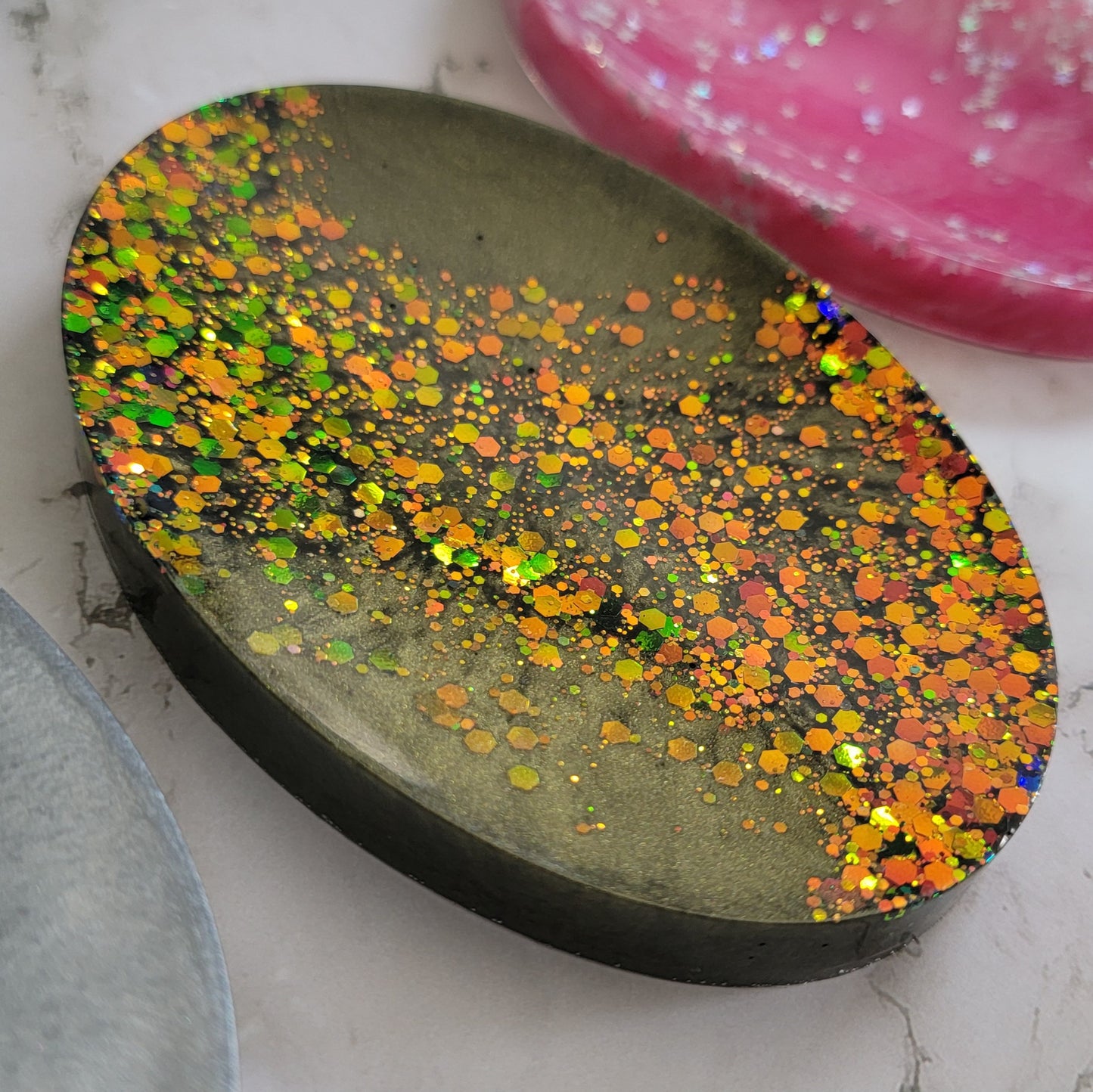 Round epoxy resin soap dish with deep olive green mica pigment and a diagonal stripe of green/gold/orange color-shift glitter.