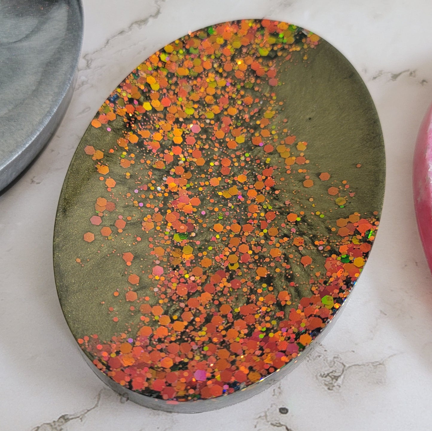 Round epoxy resin soap dish with deep olive green mica pigment and a diagonal stripe of green/gold/orange color-shift glitter.