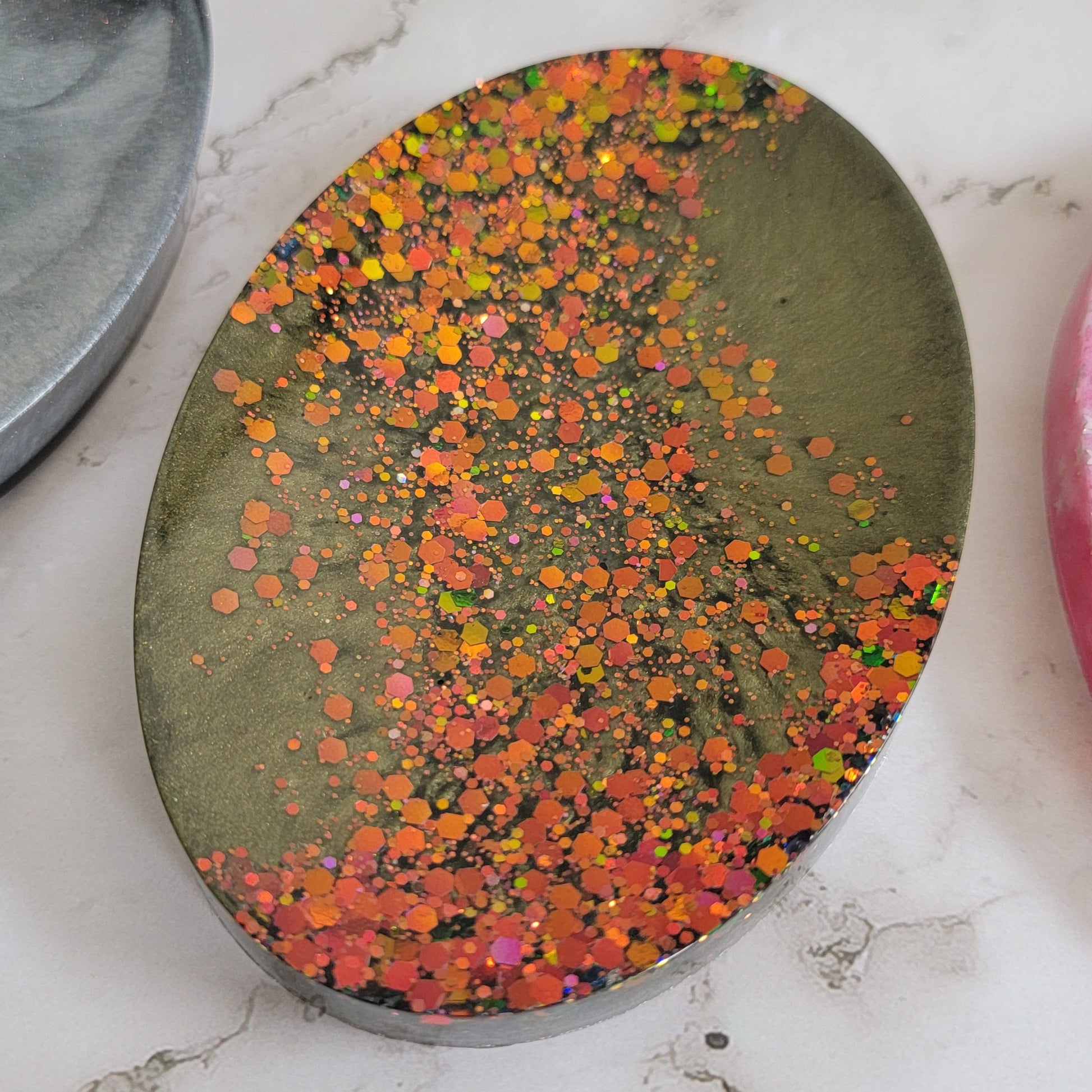 Round epoxy resin soap dish with deep olive green mica pigment and a diagonal stripe of green/gold/orange color-shift glitter.