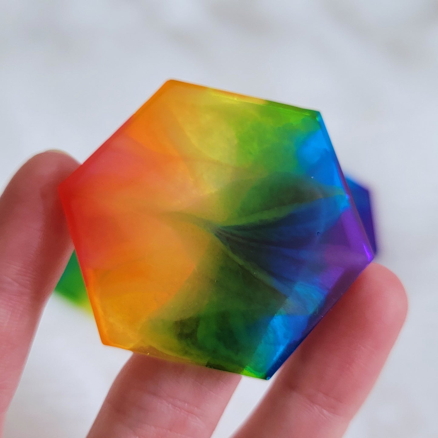 Hexagon shaped epoxy resin piece with zigzag design through translucent rainbow color stripes, being held up by a hand to show translucency..