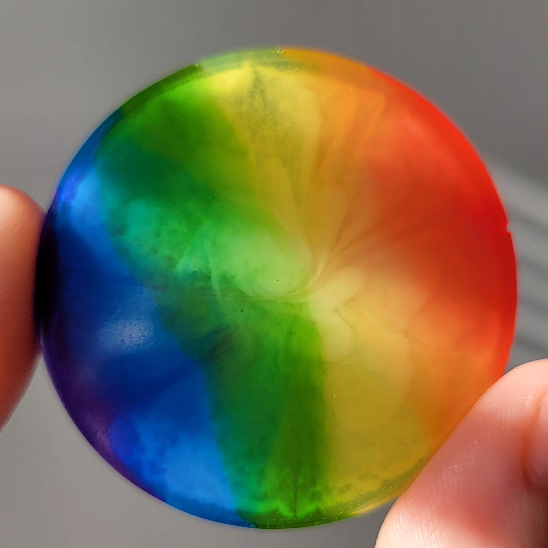 Round epoxy resin piece with subtle bloom effect in translucent rainbow stripes, being held up in light to show bloom effect.