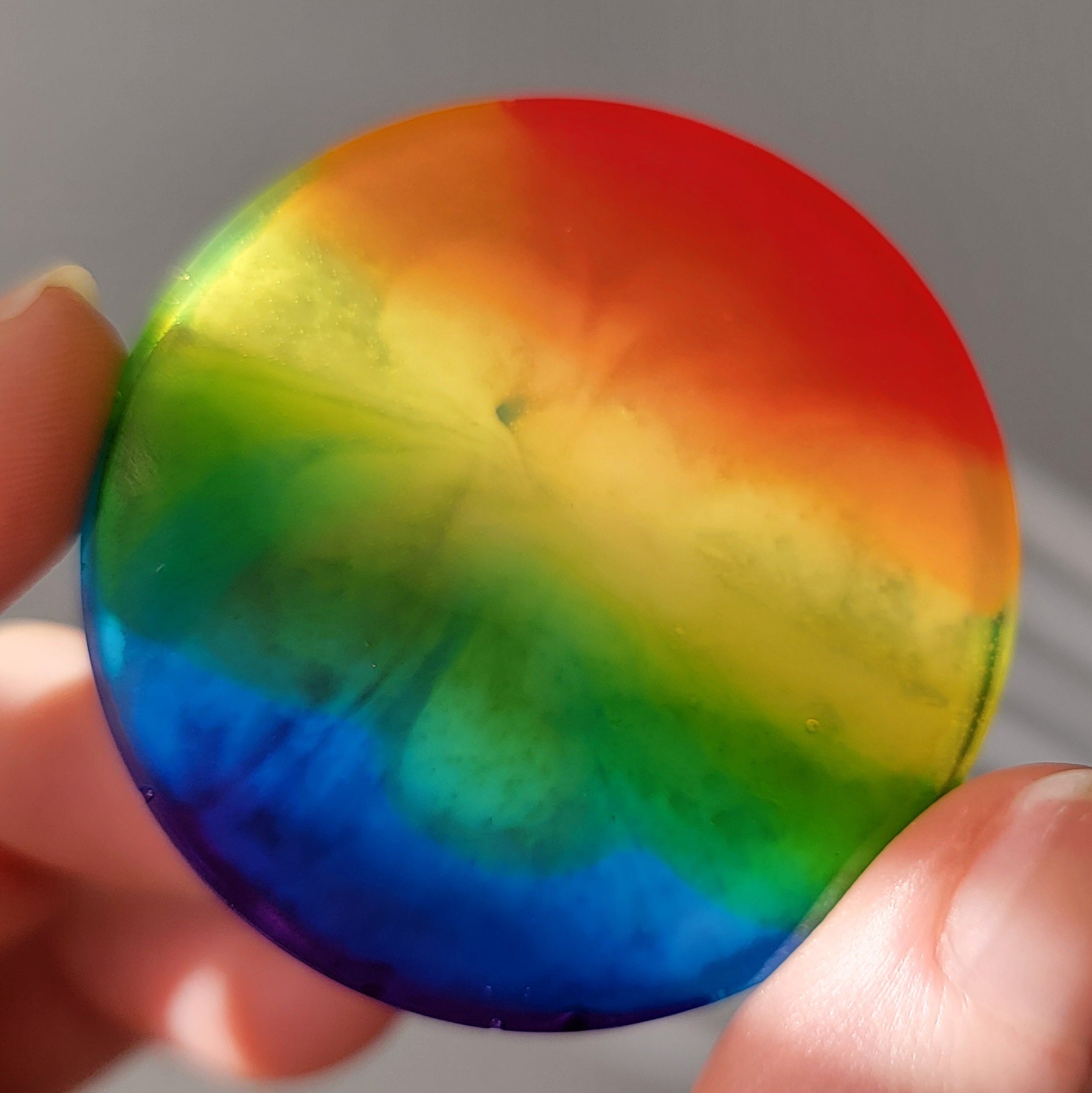 Round epoxy resin piece with subtle bloom effect in translucent rainbow stripes, being held up in light to show bloom effect.