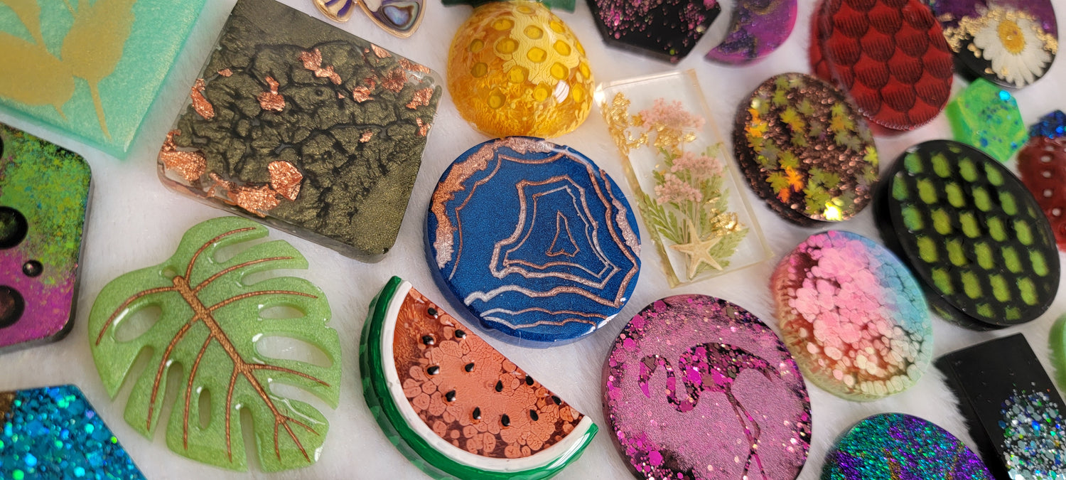 Array of epoxy resin pieces for tags keychains, styles include: sage green monstera leaf, watermelon slice, blue geode, yellow pineapple, round with hot pink flamingo design, red fish scales, floral with starfish, and various other colorful and glittery pieces.