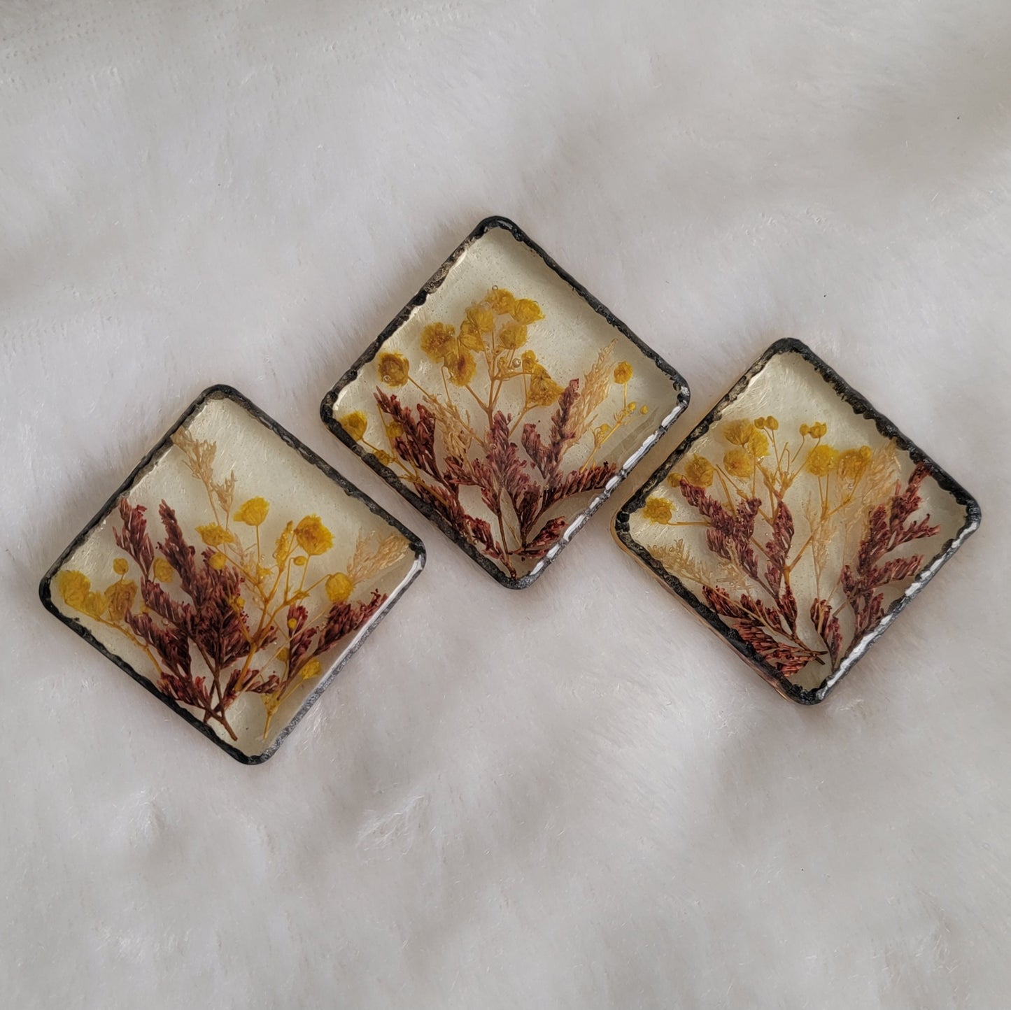 Three square epoxy resin pieces with yellow, white and red flowers cast in them and metallic black textured border.