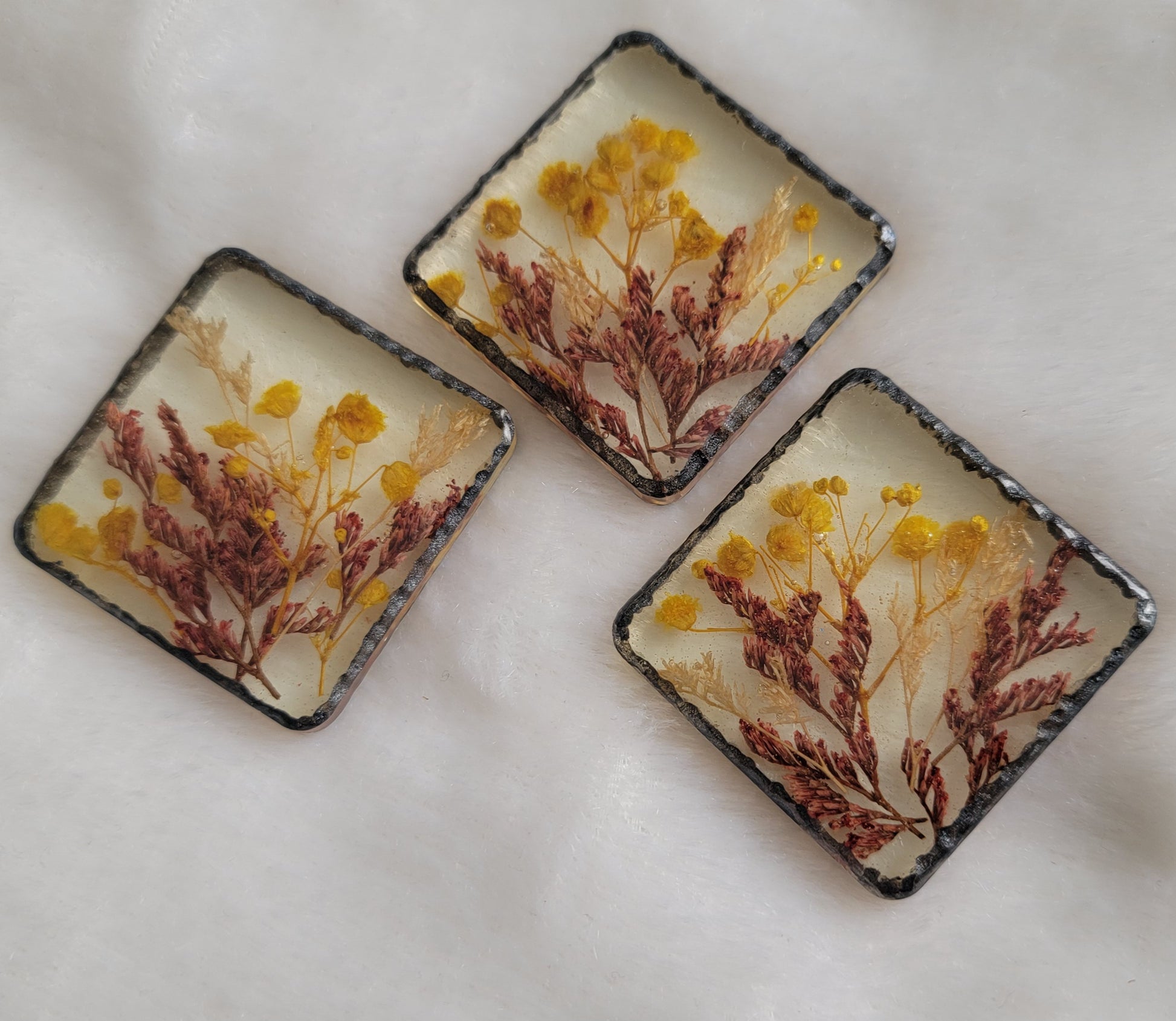 Three square epoxy resin pieces with yellow, white and red flowers cast in them and metallic black textured border.