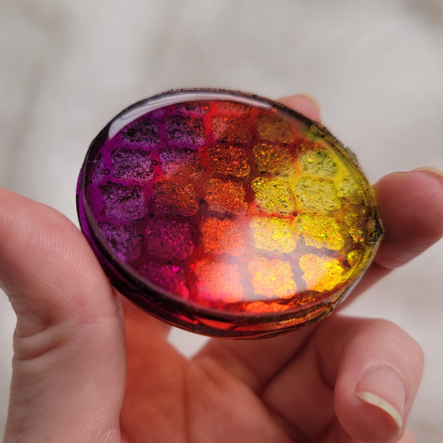 Round epoxy resin piece held in hand, translucent yellow red and purple with metallic iridescent pigment in geometric tile pattern.