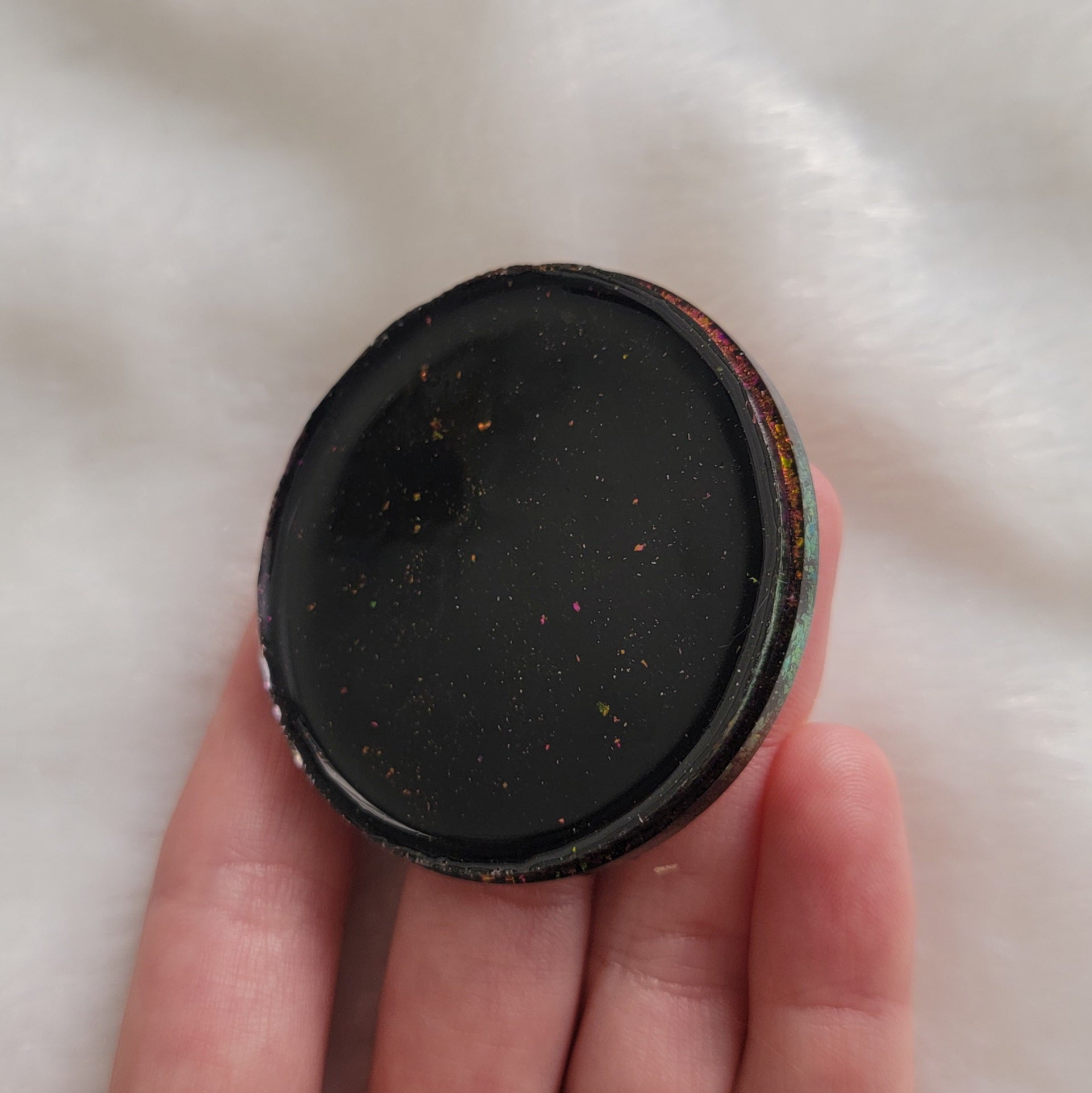 Round epoxy resin piece held in hand, black surface with some pink/gold color shift pigment flakes.