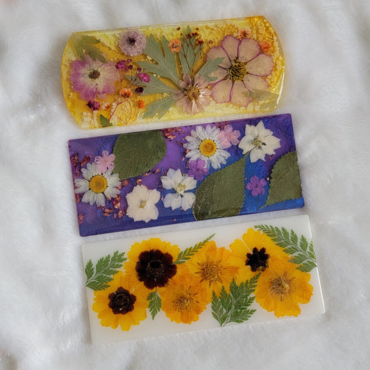 Three rectangular resin pieces. Yellow, purple, and white bases with pressed flowers and leaves.