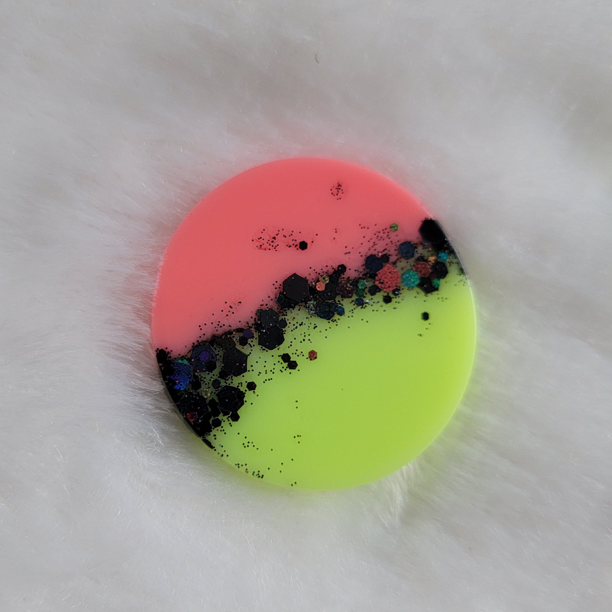 Round epoxy resin piece, half neon pink and half neon yellow-green with stripe of black holographic glitter in the center.