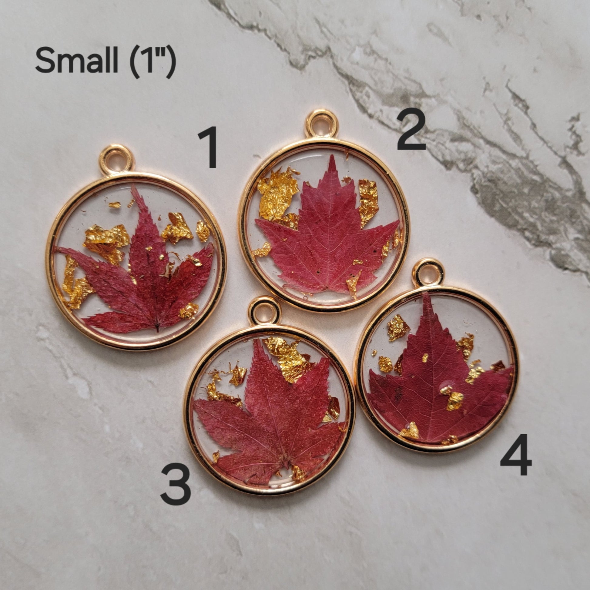 Four round epoxy resin pieces in gold tone metal frames with deep red fall leaves and gold foil flakes cast in them, photo shows options for small 1" size.