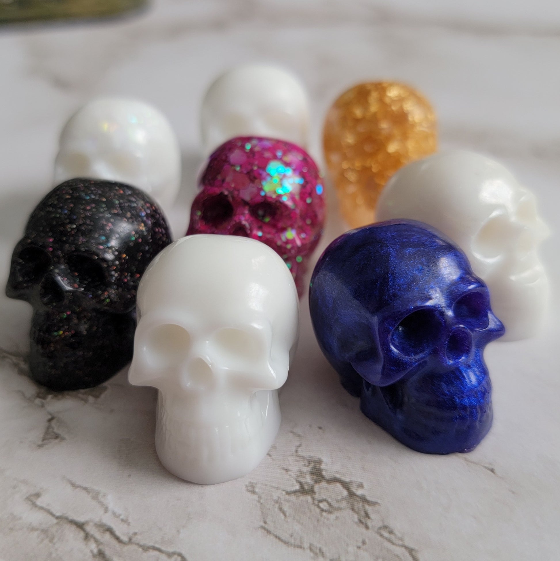 Eight small epoxy resin skulls shown in white, pink, purple, gold, and black.