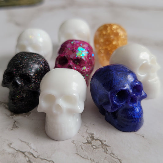 Eight small epoxy resin skulls shown in white, pink, purple, gold, and black.