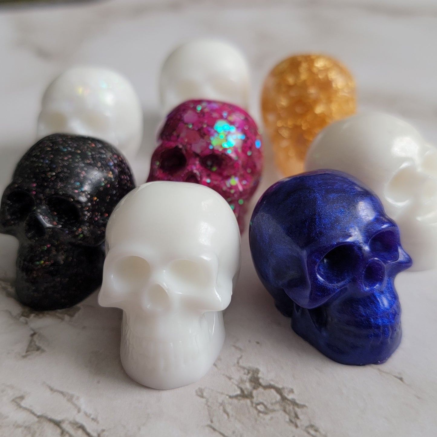 Eight small epoxy resin skulls shown in white, pink, purple, gold, and black.