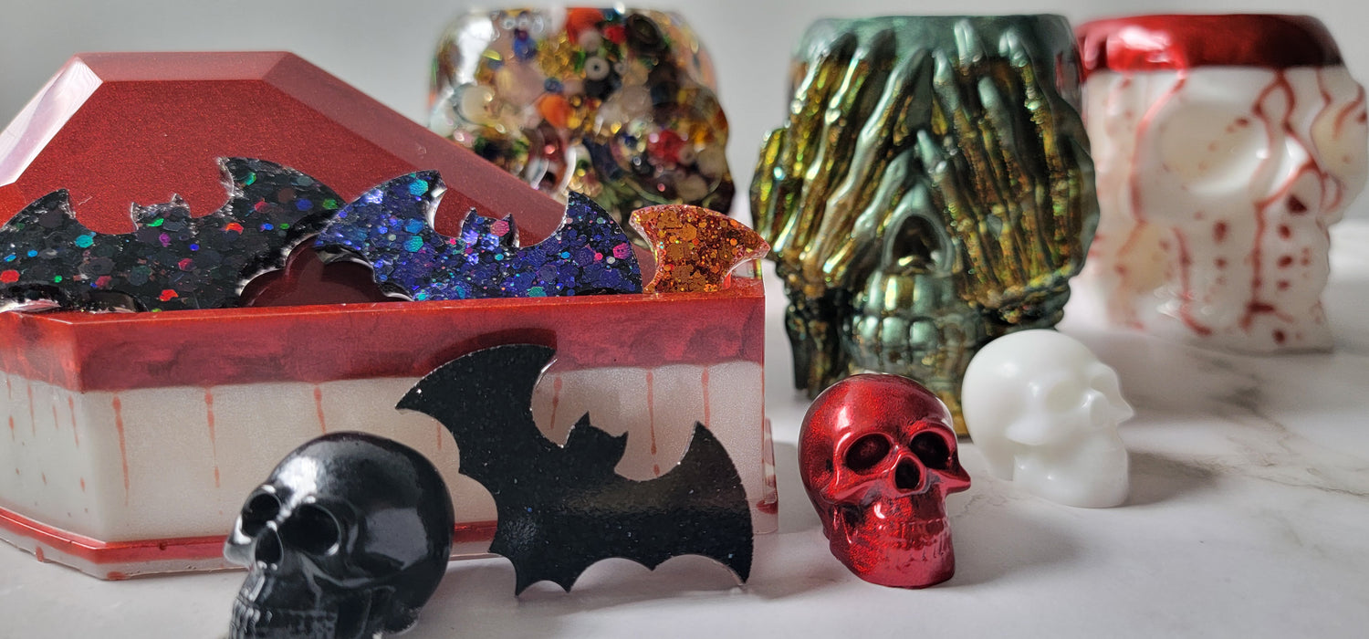 Halloween themed resin products. Small glitter bats emerge from coffin shaped trinket dish. with small skulls surrounding. Larger skull shaped plant holders in background.