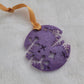 A round epoxy resin ornament that has snowflake shapes cutout from each side, made with lavender pigment and a gold ribbon attached.