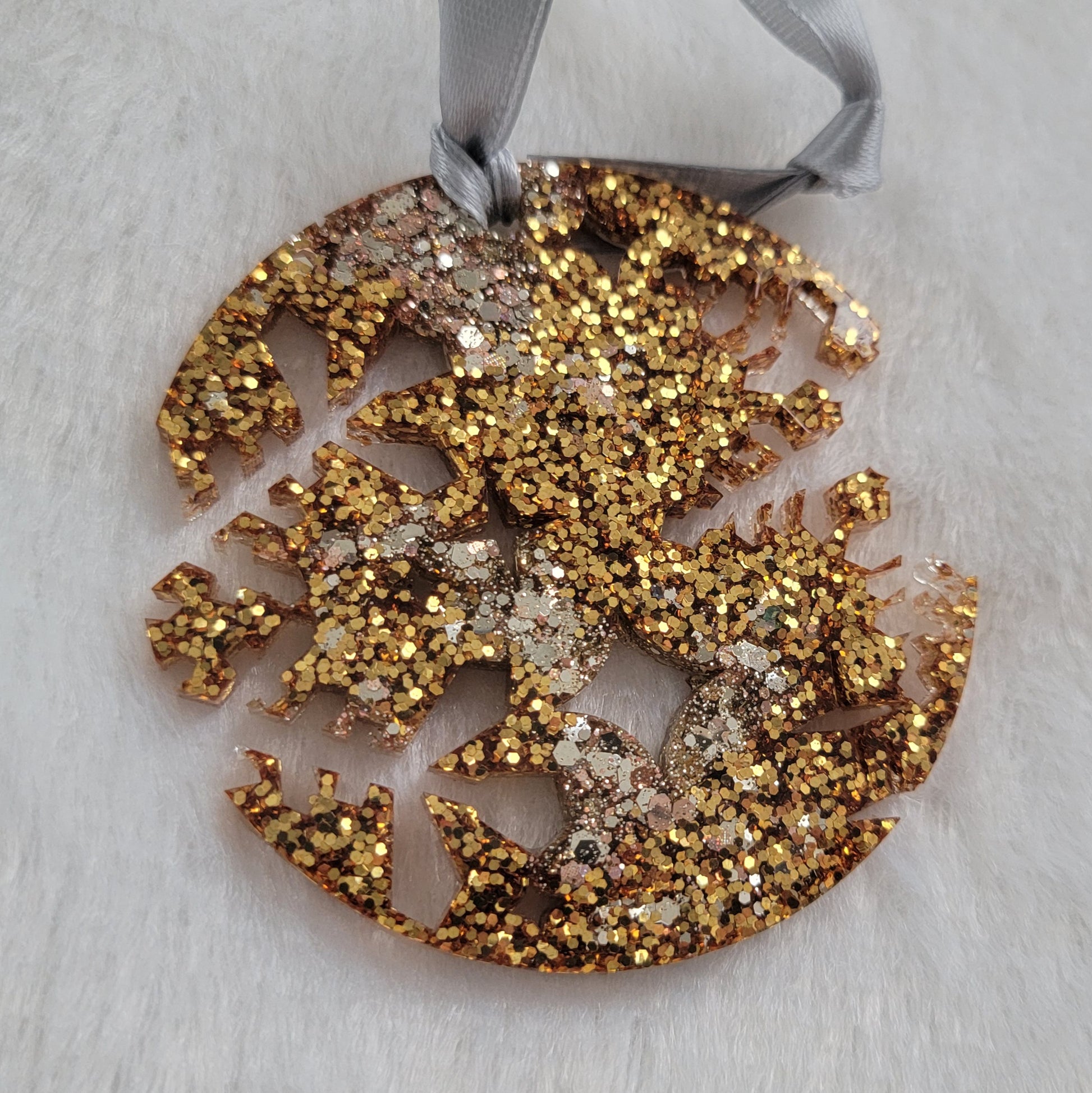 A round epoxy resin ornament that has snowflake shapes cutout from each side, made with a mix of gold, silver, and rose gold glitter and a silver ribbon attached.