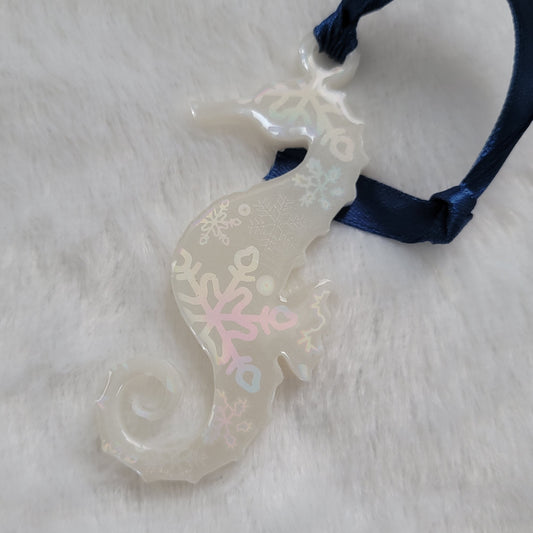 Seahorse shaped epoxy resin ornament, made with white pigment with a holographic snowflake print vinyl covering the entire piece and a dark blue ribbon attached.