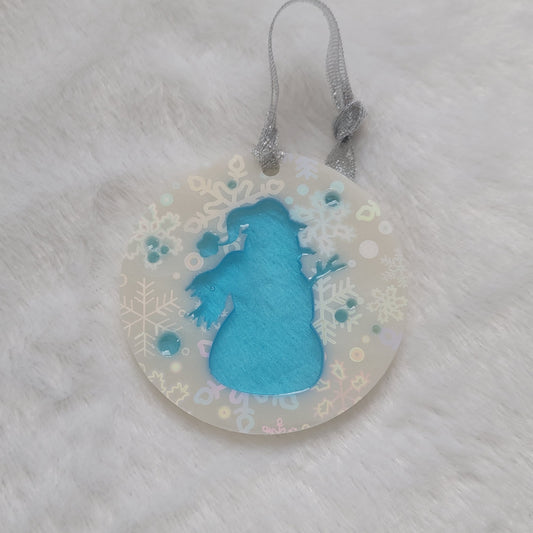 A round epoxy resin ornament that has a translucent blue snowman silhouette in the middle, made with white pigment with a holographic snowflake print vinyl covering the entire piece and a sparkling silver ribbon attached.