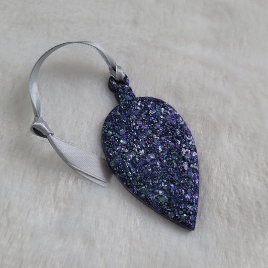 Drop shaped epoxy resin ornament that has a crystalline texture, made with purple and aqua blue holographic glitter and a silver ribbon attached.