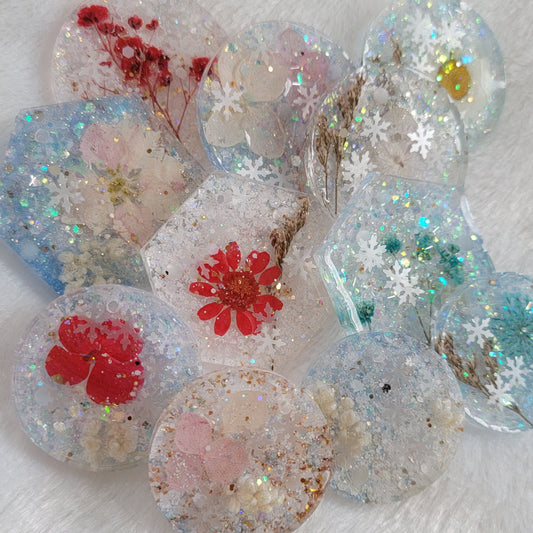 Array of round a hexagon shaped glittery epoxy resin pieces that contain snowflake glitters and pressed flowers.