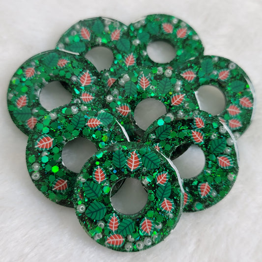 Eight small epoxy resin pieces made to look like holiday wreaths. They are wreath shaped, contain green glitter, small pearly white beads, and have green and red polymer leaves.