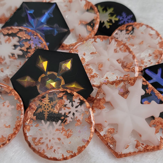 An array of epoxy resin pieces, hexagon shaped and round, containing small resin snowflake. Some with black backgrounds with iridescent snowflakes and some transparent with metallic copper accents and white snowflakes.