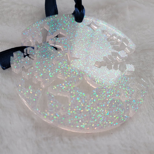 A round epoxy resin ornament that has snowflake shapes cutout from each side, made with iridescent white glitter and a dark blue ribbon attached.