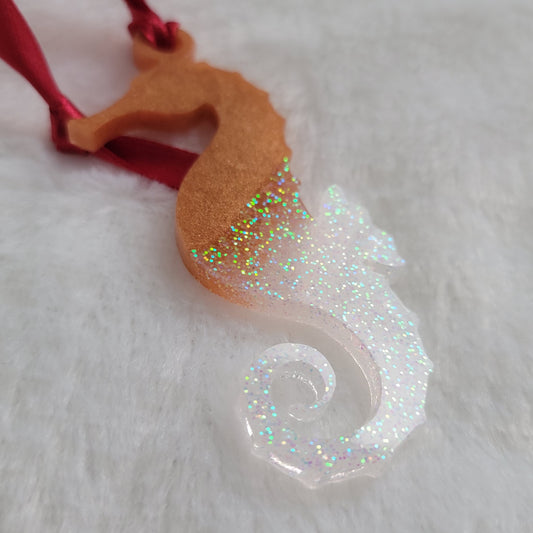 Seahorse shaped epoxy resin ornament, made with golden bronze pigment and iridescent white glitter and a red ribbon attached.