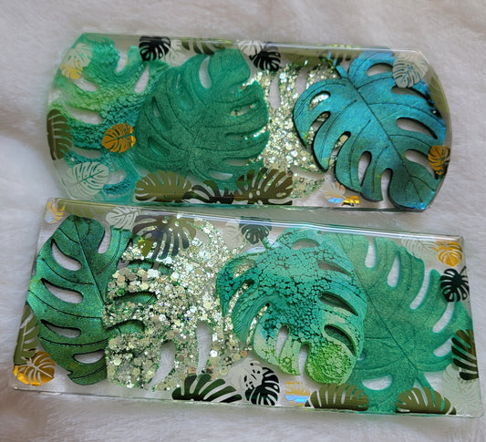 Two rectangular epoxy resin pieces for crate tags or home decor, one with rounded edges and one with rectangular. Each has four large resin monstera leaves in varied green pigments and glitter, and several smaller vinyl monstera leaves in shades of green and gold.