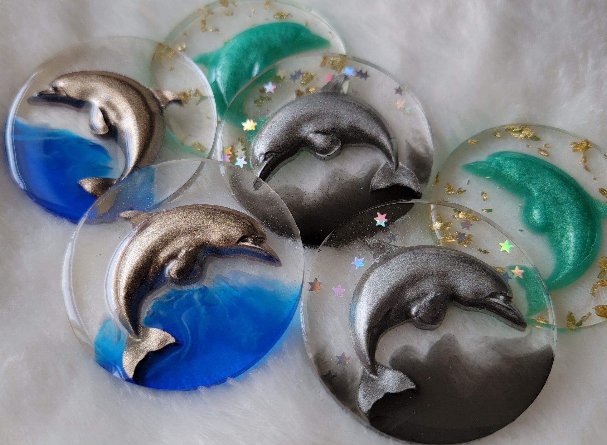 Six round epoxy resin pieces, each contains a three dimensional resin cast dolphin - two warm silver with bright blue waves, two cool silver with holographic stars and black waves, and two aqua with gold foil flakes.