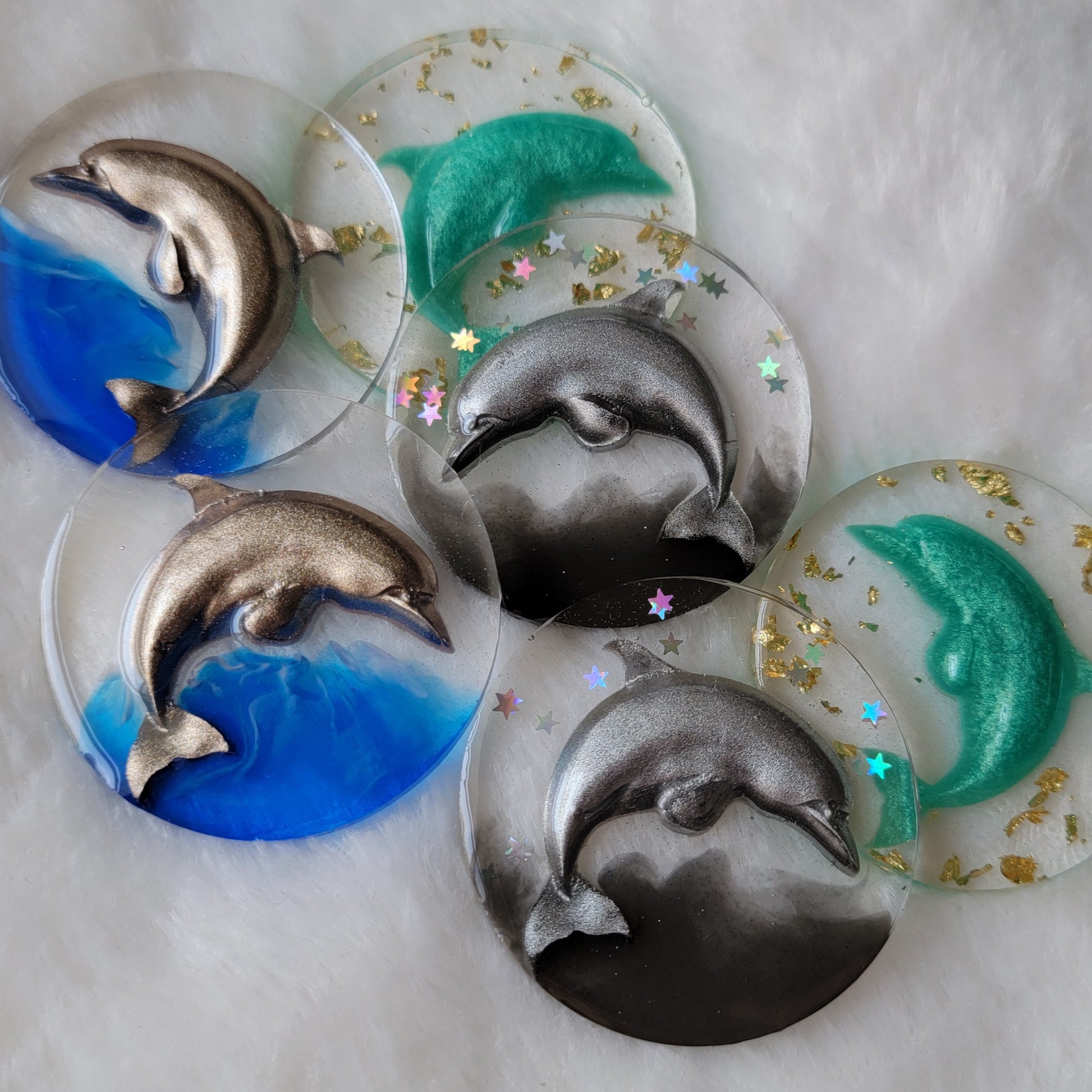 Six round epoxy resin pieces, each contains a three dimensional resin cast dolphin - two warm silver with bright blue waves, two cool silver with holographic stars and black waves, and two aqua with gold foil flakes.