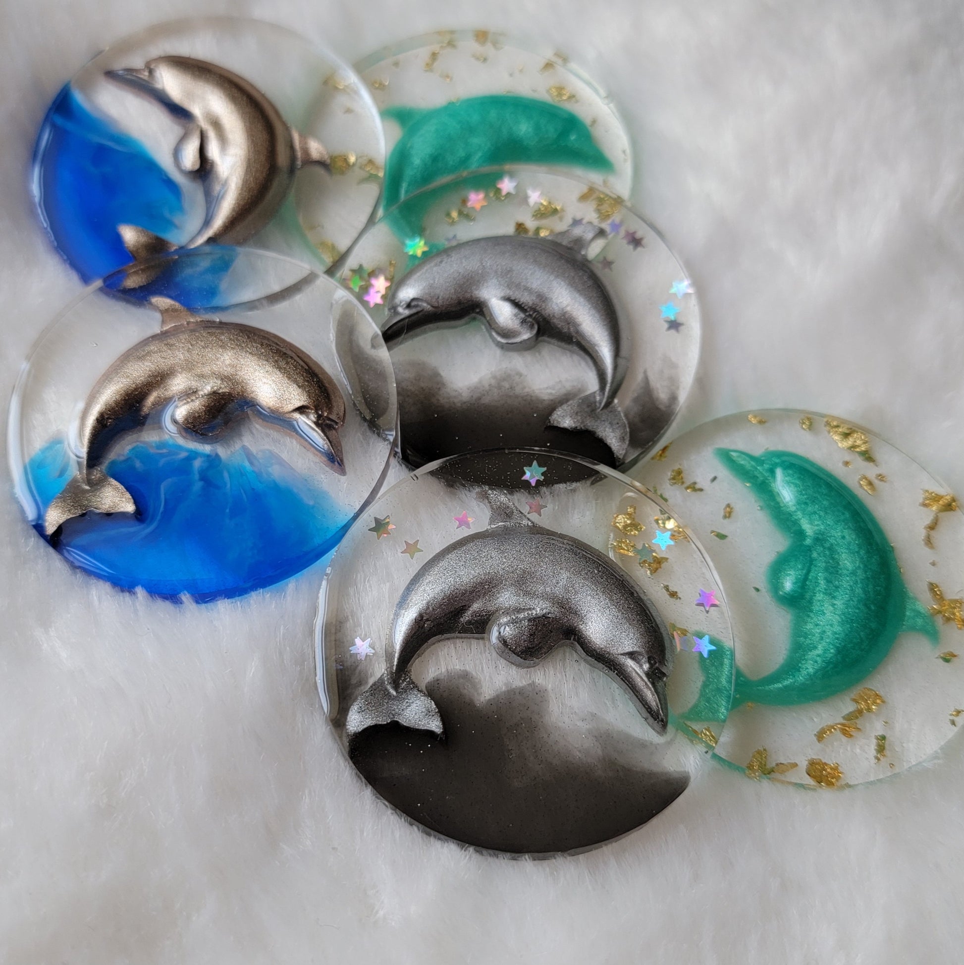 Six round epoxy resin pieces, each contains a three dimensional resin cast dolphin - two warm silver with bright blue waves, two cool silver with holographic stars and black waves, and two aqua with gold foil flakes.