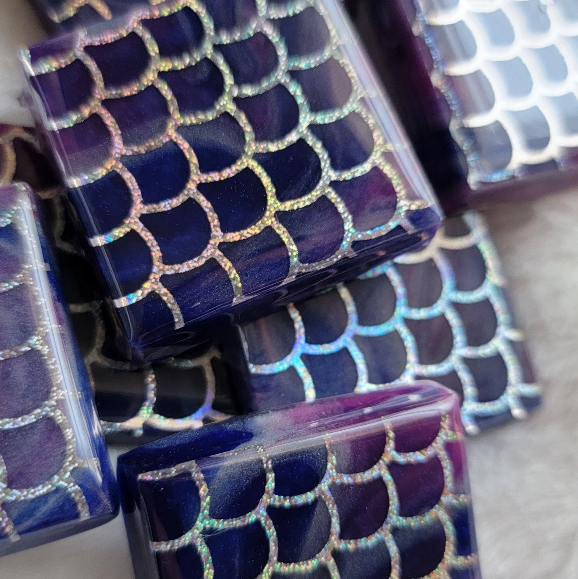 Close up of square epoxy resin pieces, blue and purple swirled bases with holographic silver fish scale design in vinyl.