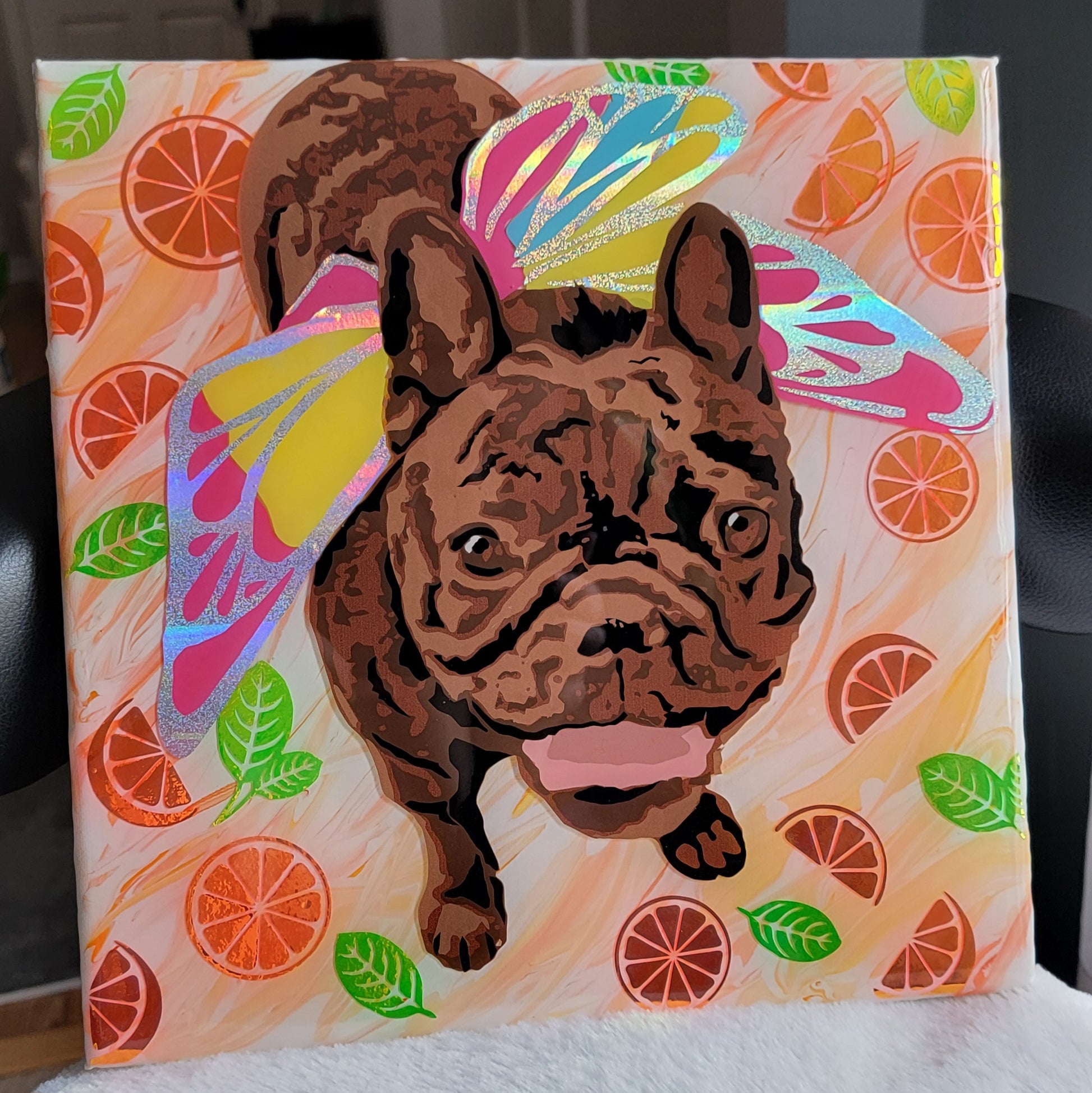 Portrait of a brown brindle French bulldog wearing rainbow fairy wings made with layers of adhesive vinyl on a canvas. The background has orange swirled acrylic paint and is adorned with shiny vinyl orange slices and leaves.
