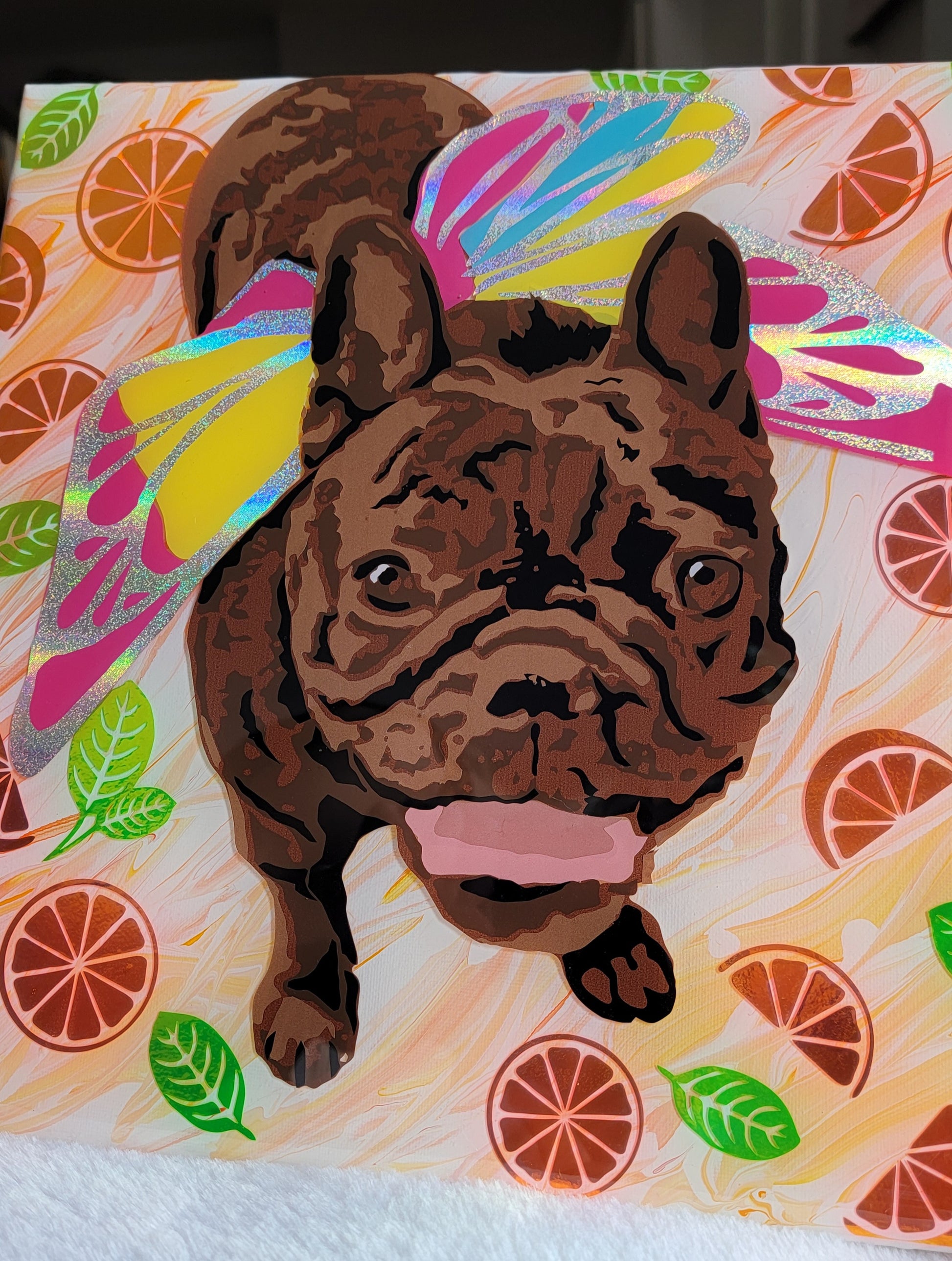 Portrait of a brown brindle French bulldog wearing rainbow fairy wings made with layers of adhesive vinyl on a canvas. The background has orange swirled acrylic paint and is adorned with shiny vinyl orange slices and leaves.